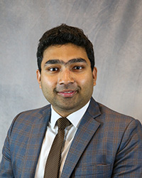 Headshot of Adil Mohammed wearing a blue checkered blazer, white shirt and brown tie.
