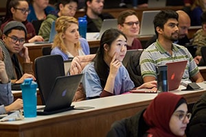 A young Asian woman puts her finger on her cheek and her other hand on her laptop as she looks forward in a lecture hall.