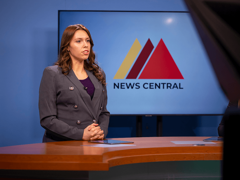 A woman with long brown hair stands at a news desk in front of a screen with the News Central icon on it.