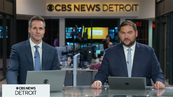 Two male new anchors from CBS New Detroit sitting at a glass table, wearing blue suits with laptops in front of them.