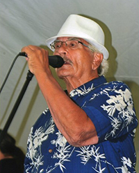 Image of Larry T. Reynolds wearing a white hat and blue and white shirt while speaking into a microphone.