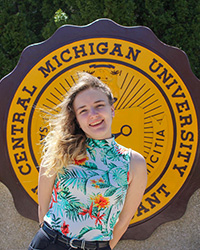 Jessica Reinhart in a floral shirt standing in front of the CMU seal, smiling at the camera.