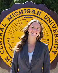 Kelsey Muth in a grey suit and white top standing in front of the CMU seal smiling at the camera.