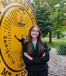 Lauren Day in a dark suit and green top standing in front of the CMU seal smiling at the camera.