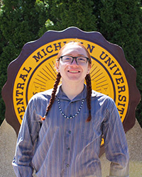 Mason Squillets'Peterson in a striped long sleeve shirt and glasses, standing in front of the CMU seal, smiling at the camera.
