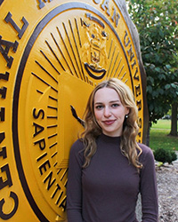 Tiffany Jurge in a dark long sleeve top standing in front of the CMU seal smiling at the camera.