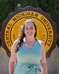 Zoey Archbold in a pale green dress, standing in front of the CMU seal, smiling at the camera.