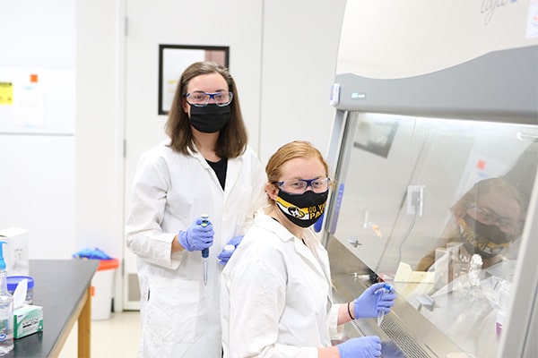Two students in a research lab wearing white lab coats, masks, gloves, and eye protection and holding syringes look at the camera.