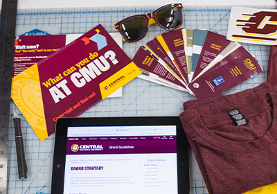 Central Michigan University postcard with blue, gold and red chevrons, several maroon brochures, maroon and gold sunglass and an ipad lay on a light tan tabletop with a grid pattern.