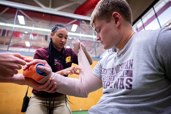 A female sports medicine student wearing a maroon CMU shirt splints and wraps the arm of a male student wearing a gray t-shirt.