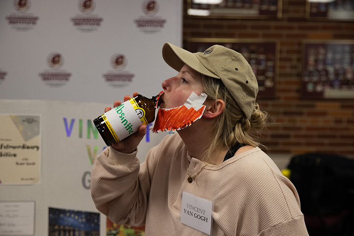 A female CMU student dresses up as Vincent Van Gogh, wearing a light brown shirt and hat, wearing a beard made out of a paper plate, and drinking a fake bottle of absinthe.