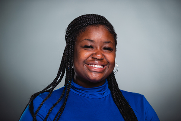 Professional headshot of a smiling Anyah Lewis wearing a blue shirt against a light grey background.