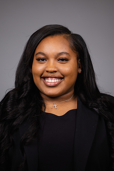 Professional headshot of a smiling Zayna Goode wearing a black jacket and black shirt while looking at the camera.