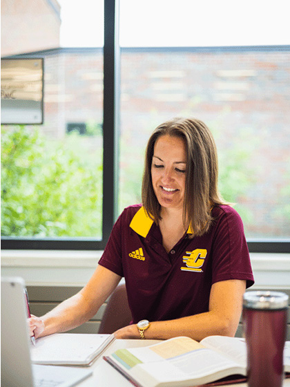Online-Campus-Admissions-480x720 Woman in burgandy and yellow CMU t-shirt writing in a notebook with a laptop, textbook and a tumbler on a desk