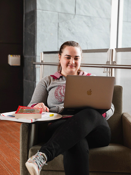 Online-Campus-Military-and-Veterans-480x720 A woman in a grey CMU hoody siting on a green armchair with books and a laptop