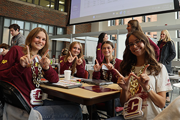 Four ERPsim participants smile and make CMU hand signs at a table with laptops, wearing gold chains with CMU emblems.
