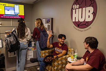 Two students sit in chairs, while two stand and speak to an Ambassador at the desk in the HUB.