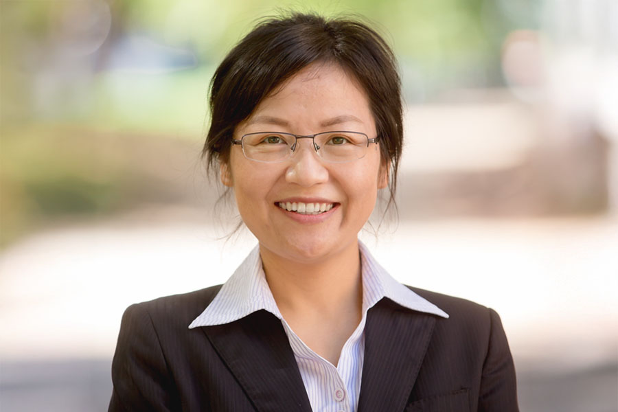 900x600_Article_Inset_Template Li Zhang stands outdoors and faces the camera and smiles. She has black hair that is pulled back. She wears wire frame glasses. She has on a black blazer and a white button down shirt with dark pinstripes.