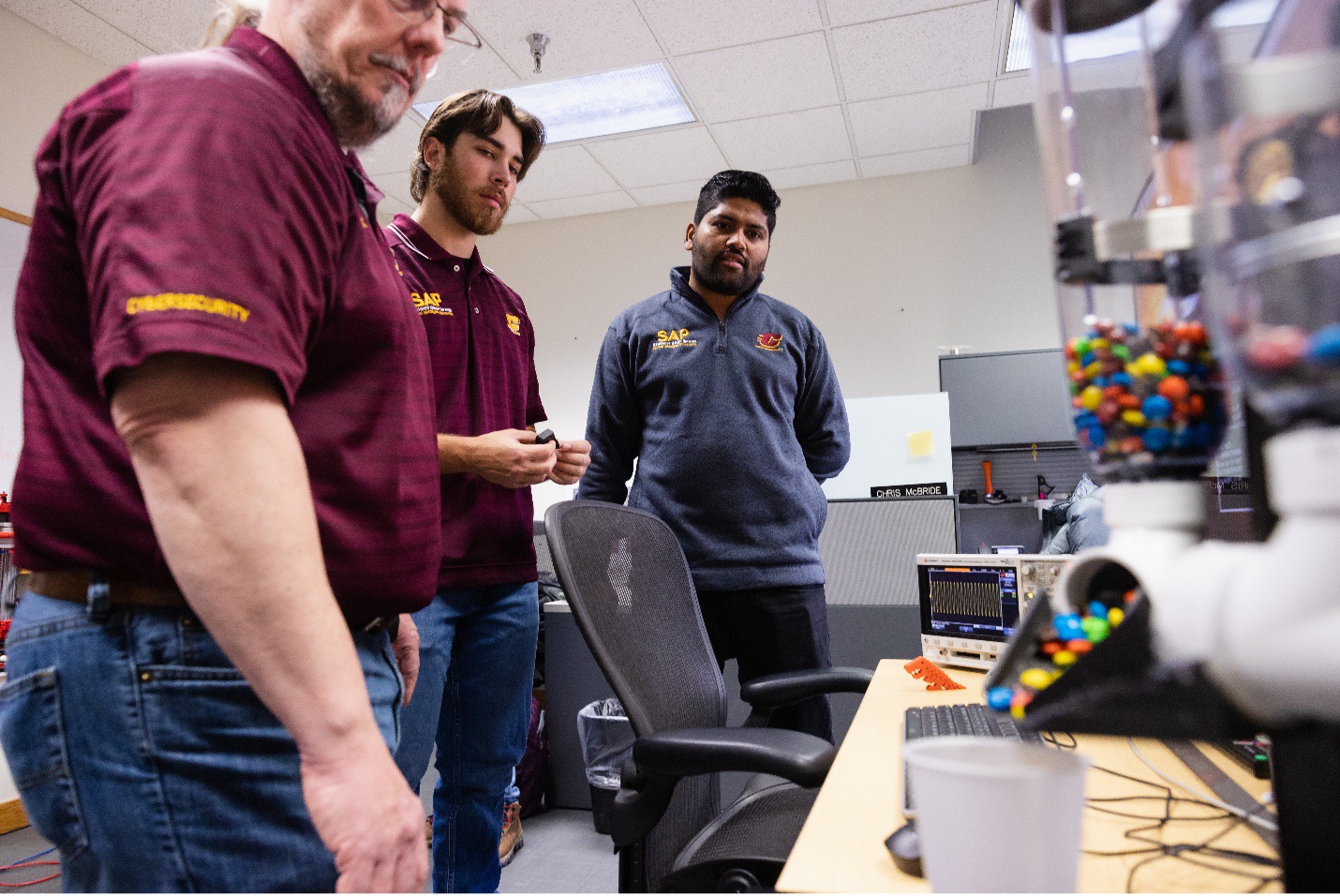 2022-325-0054-CBA VMD SAP lab as.jpg Three men in CMU t-shirts standing in front of a desk with a candy machine