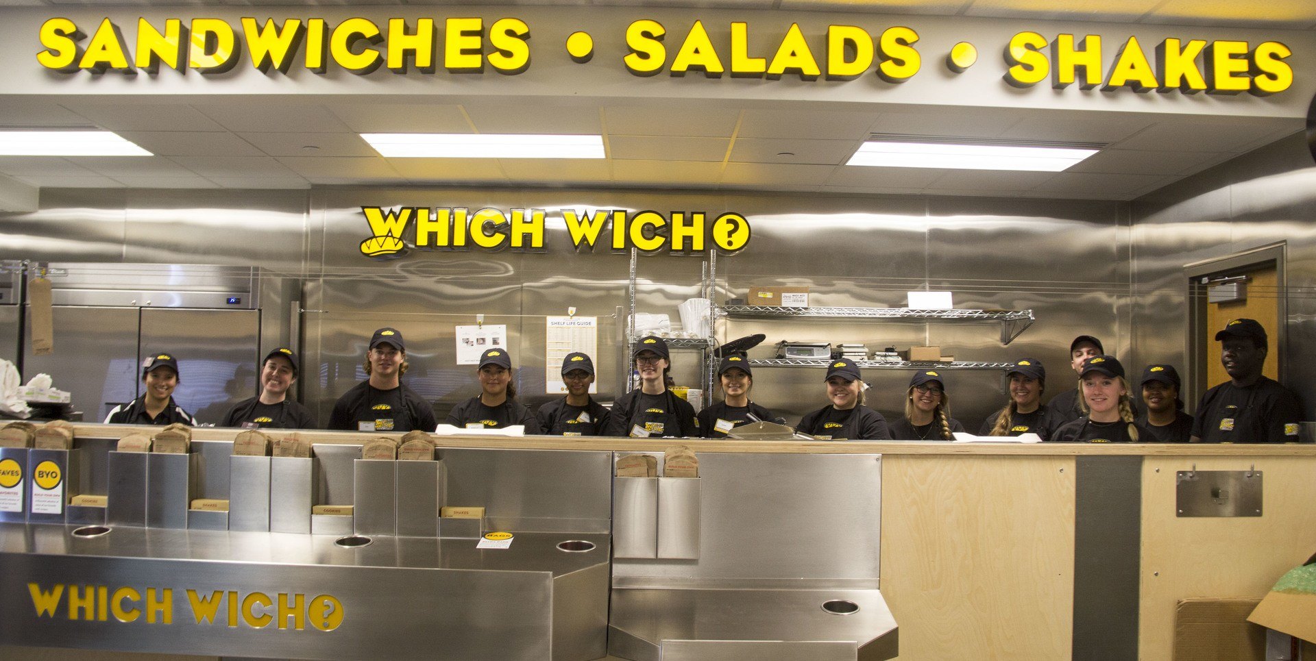 Young adults are lined up behind the counter of the sandwhich shop Which Wich.