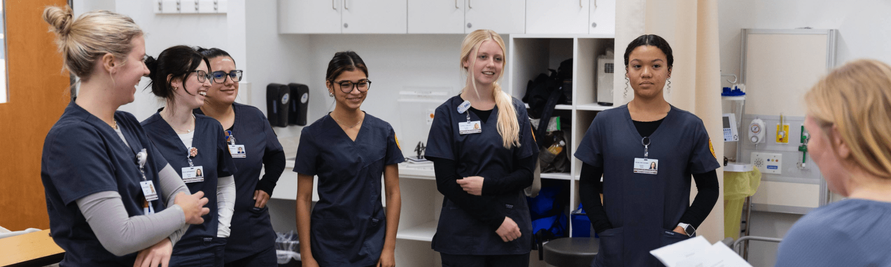 Nursing students in navy scrubs listen to an instructor in a clinical skills lab.