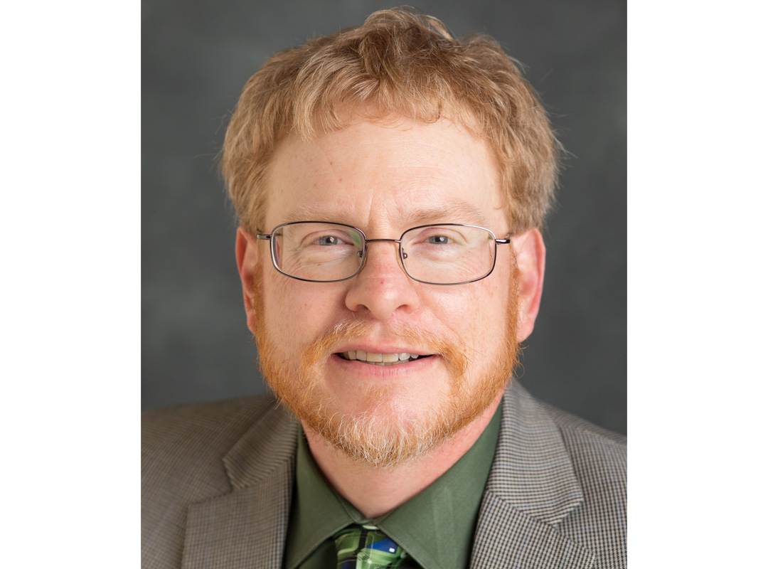 Derek H. Alderman wears a green shirt, tie and blazer in a professional headshot.
