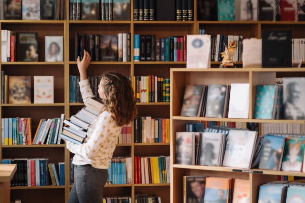 A librarian from a public library holding a stack of books while reaching up to retrieve another. In the foreground and background are shelves of books.