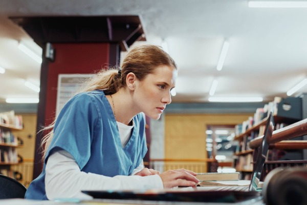 Med student in the library researching on a laptop. the student is wearing blue medical scrubs.