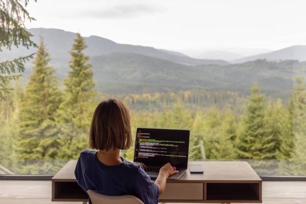 a student sitting at a desk working on a laptop in fromt of a large window. Out this window is a view of mountains and pine trees.