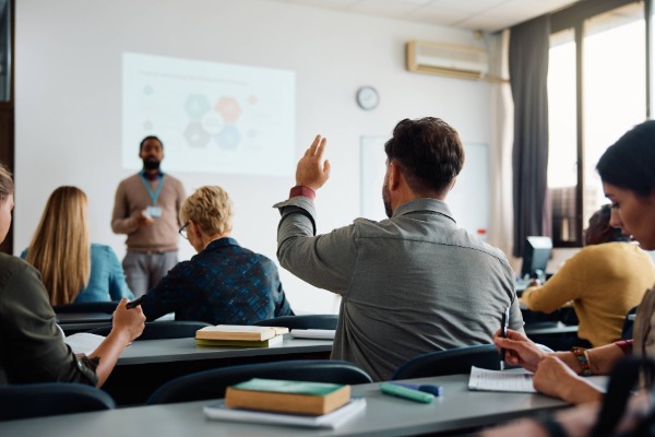 A student raising his hand in a college class, in the background is the professor who is calling on him to answer.