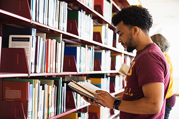 A student wearing a maroon shirt browses through books in the library stacks.