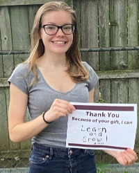 a smiling woman holding a sign that says, Thank you. Because of your gift, I can learn and grow