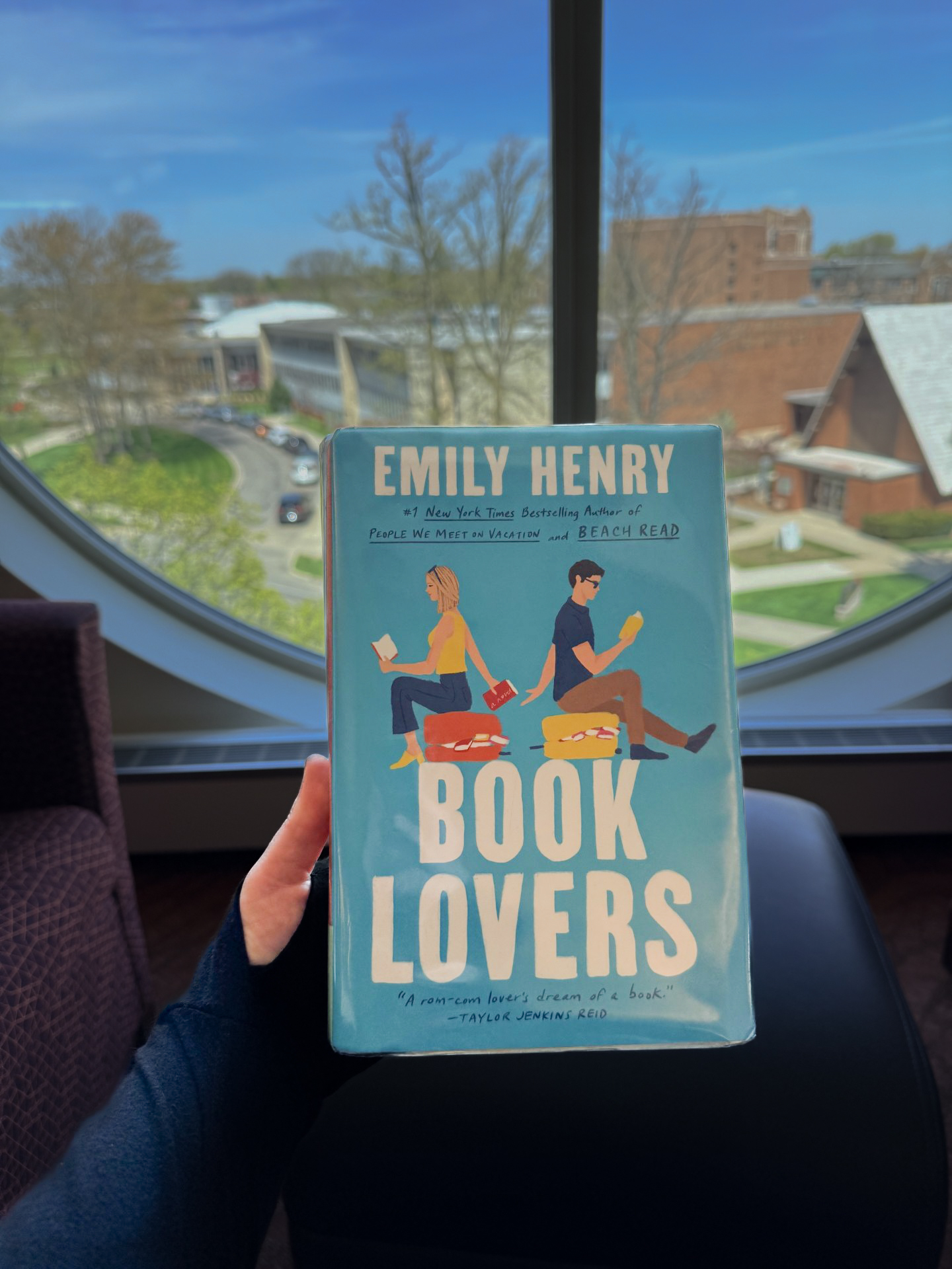 Browsing collection book cover Person's hand holding Book Lovers by Emily Henry in front of a library round window. The cover features two people reading books facing away from each other, but passing a book behind their backs.