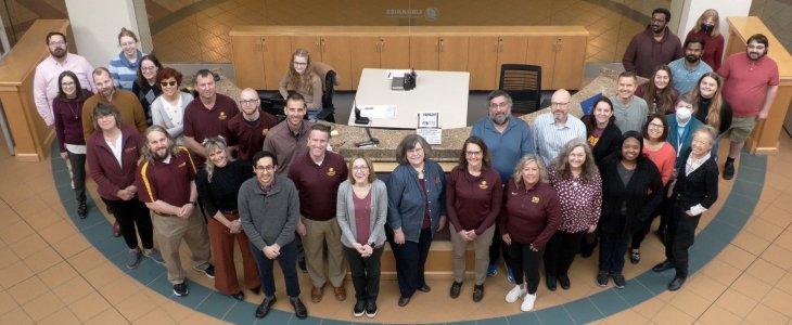Photo of Library Faculty and Staff standing in front of the first floor info desk. Photo taken on a downward angle from the second floor.