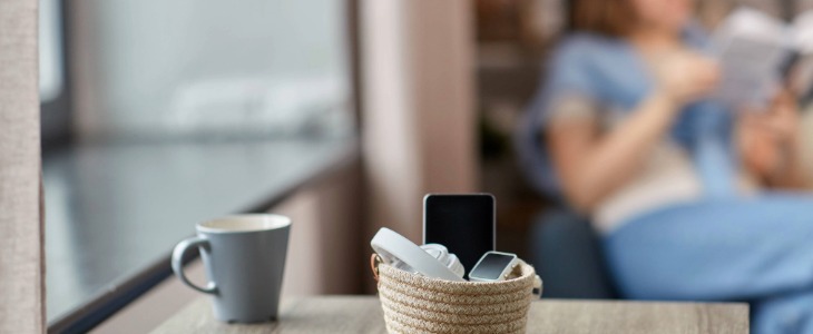Basket of electronics on a table next to a mug with a person across the room In the background reading a book.