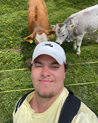 Headshot of Joseph Dinkel in a yellow shirt, white baseball cap, black backpack against a green grass pasture background with one brown and  one white gray cow.