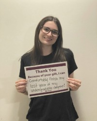 A woman holding a sign that says, Thank You, Because of your gift I can comfortably finish my last year of my undergraduate degree!