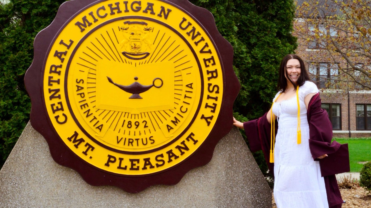 A student with cap and gown standing next to the university seal.