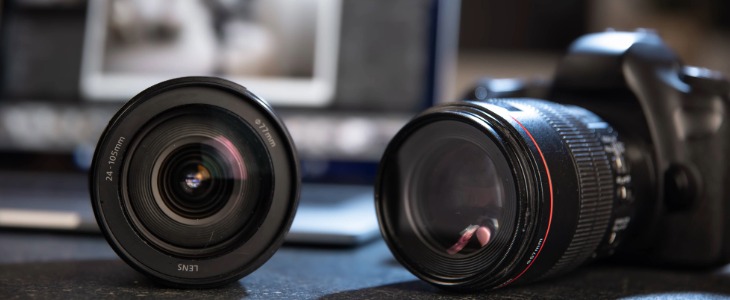 Image of a professional camera on the right and a camera lens on the left with a blurry laptop computer in the background.