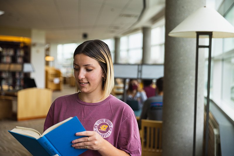 student wearing maroon and gold holds open a book and smiles while standing in the CMU Park Library