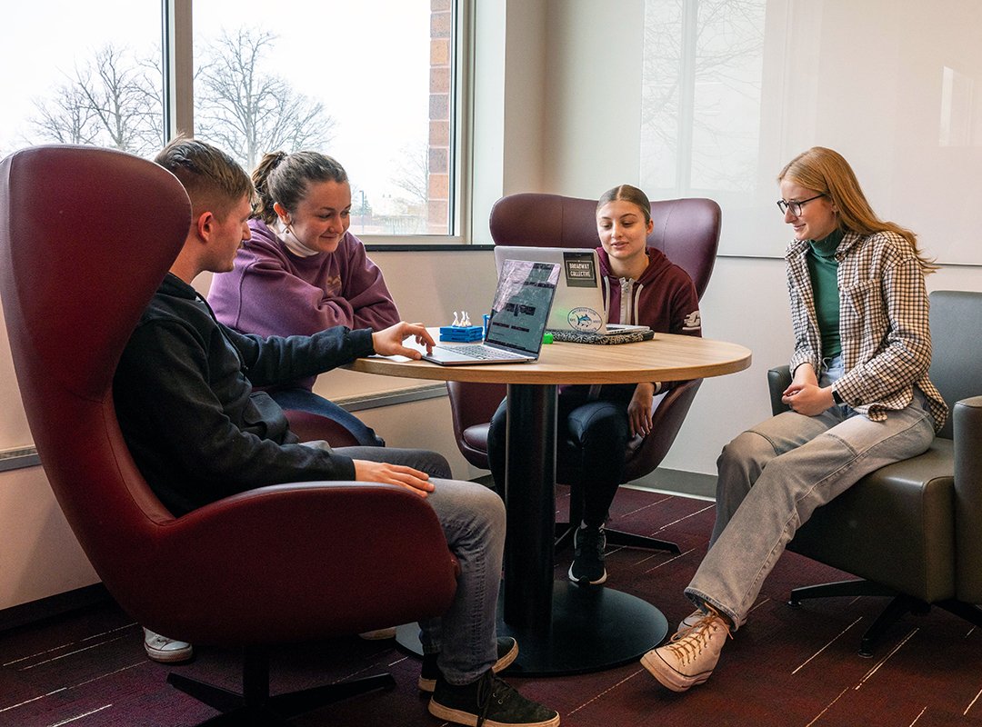 Four students sitting around a table in the Adobe Digital Lounge working at laptops and collaborating.