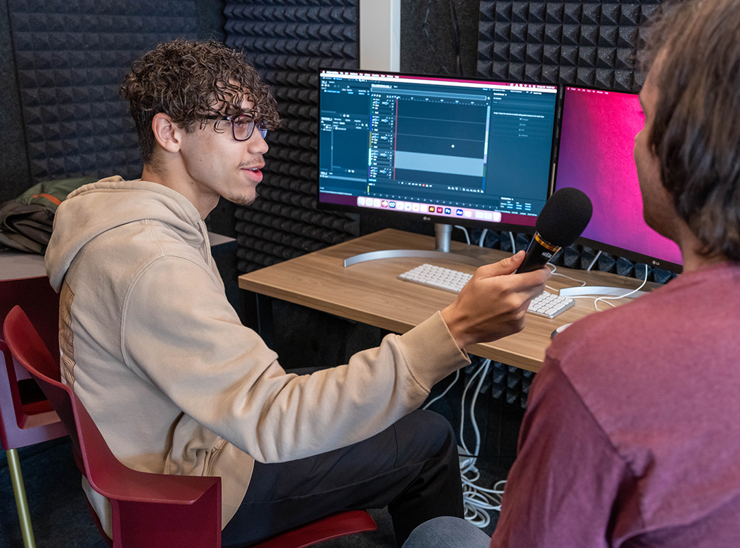 A student interviewing another student while holding a microphone in the sound recording booth in the Adobe Digital Lounge