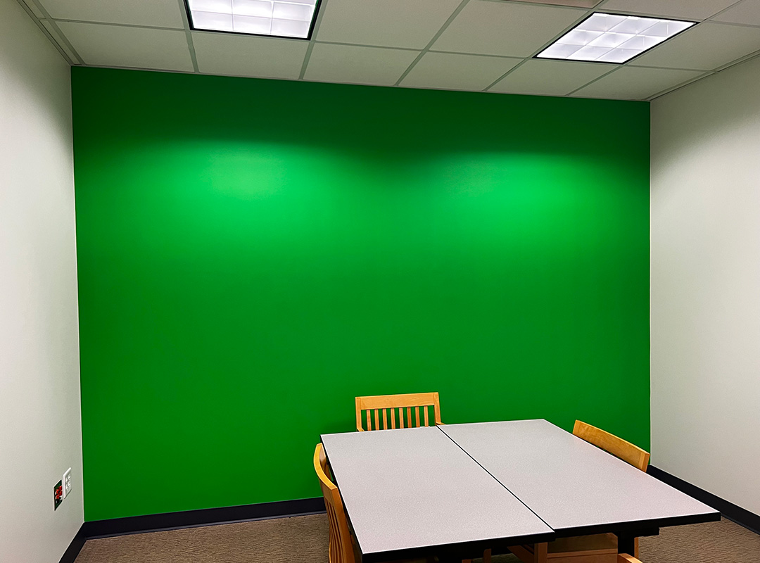 A photo of the green back wall of the green screen room that students can book for use along with the table and chairs included in the room.