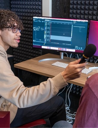 two students in the recording booth. One is holding a mic to the other interviewing them. There are two screen monitors on a desk in front of them.