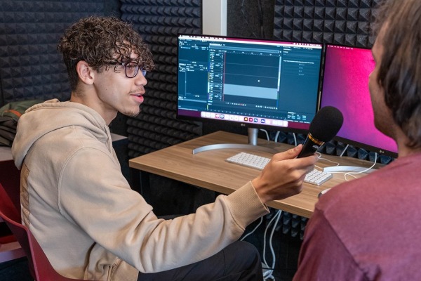 two students in the recording booth. One is holding a mic to the other interviewing them. There are two screen monitors on a desk in front of them.