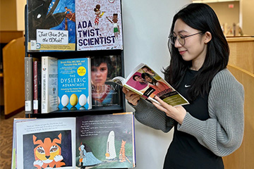 A female student with dark hair, dress, sweater, and glasses reads a book from an Autism and Neurodiversity Acceptance Month book display at CMU Libraries.