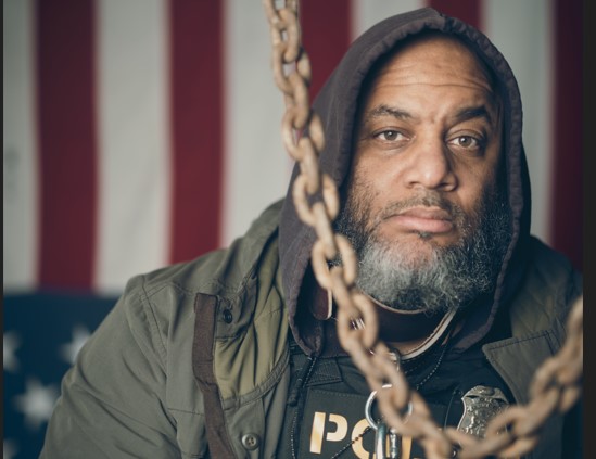 photographic art depicting man with chains in a police flack jacket in front of an American flag