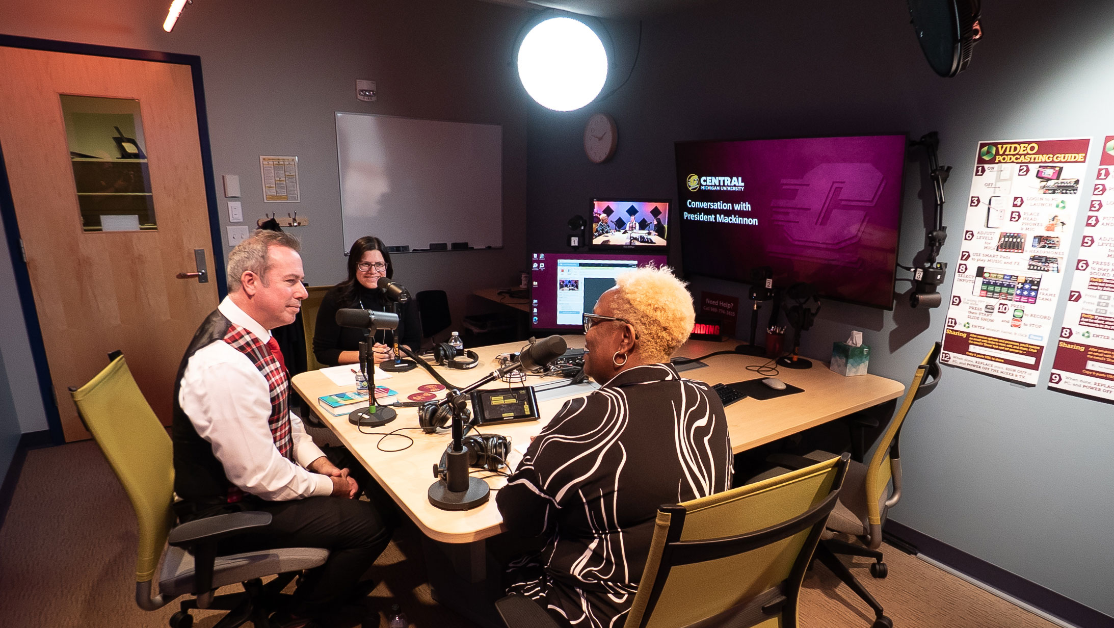 CIS-Podcast-Studio_2026_web Podcast studio wide view showing three individuals seated at a table, using the TV, mics, sound mixer, and lights.