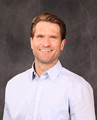 Kyle Scherr portrait, showing head and shoulders, smiling, on a gray background