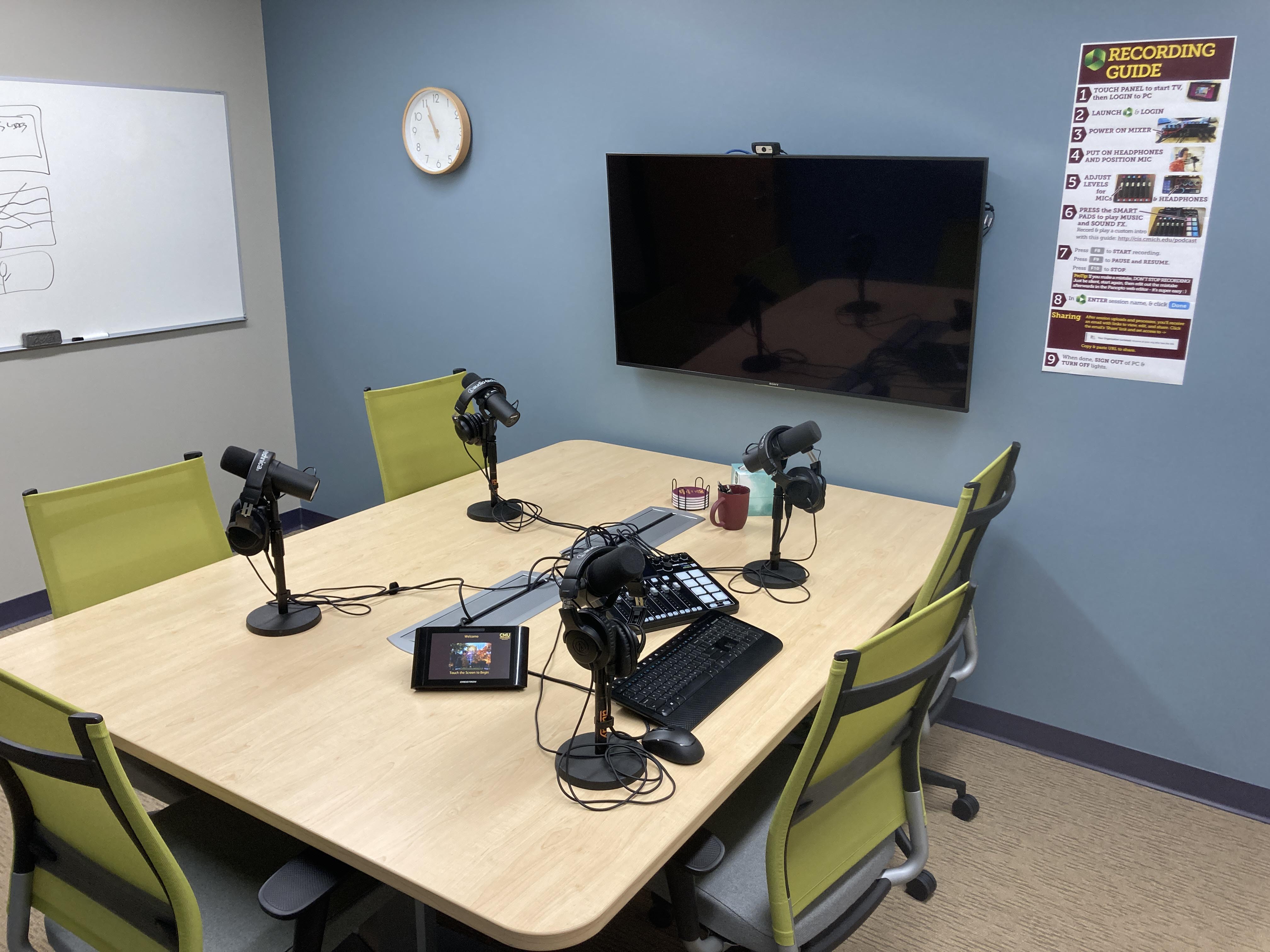 Podcast studio wide view showing TV, mics, sound mixer, table and chairs.